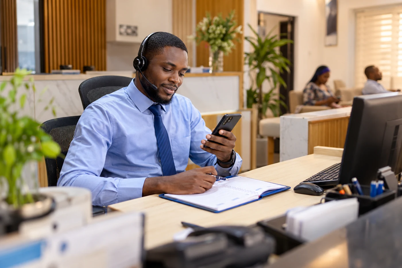 Male receptionist wearing a headset and responding to a smartphone beside a blue appointment diary.